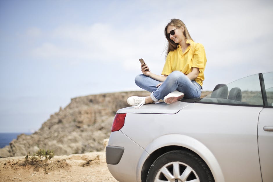 Smiling young woman sitting on a car at the beach in Mgarr, Malta using a smartphone